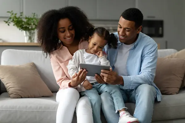 Happy black parents and child daughter using digital tablet browsing internet and watching cartoons online sitting on sofa at home