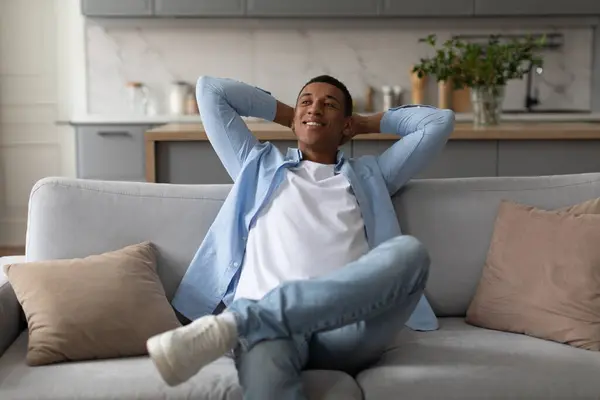Relaxed black man leaning back on couch, sitting on comfortable sofa with hands behind head, resting in cozy living room, copy space