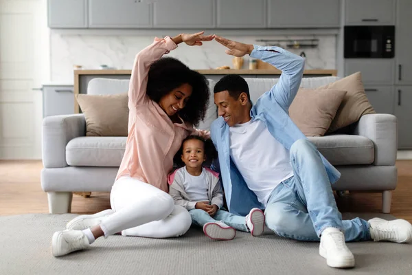Young black parents making roof symbol of hands above their little daughter, sitting on floor at home, dad and child enjoying new house