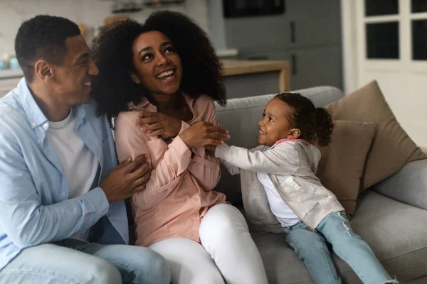 Happy black family laughs and giggles together as daughter tickles mother for fun, sitting on sofa at home
