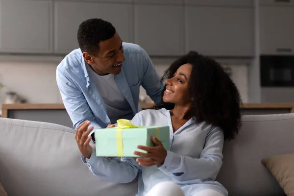 Black husband giving wrapped gift to wife surprising her on valentines day sitting on sofa at home. Family holidays celebration concept