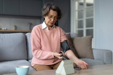 Üst düzey bir kadın, tansiyon ölçümlerini kontrol etmek için tıbbi cihaz kullanıyor. Kanepede oturup yüksek tansiyondan muzdarip, sağlığıyla ilgileniyor.