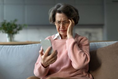 Depresyondaki yaşlı kadın telefon ekranına bakıyor ve kafaya dokunuyor, oturma odasında koltukta oturuyor, mesajdaki kötü haberleri okuyor, cihazla ilgili bir sorun var.