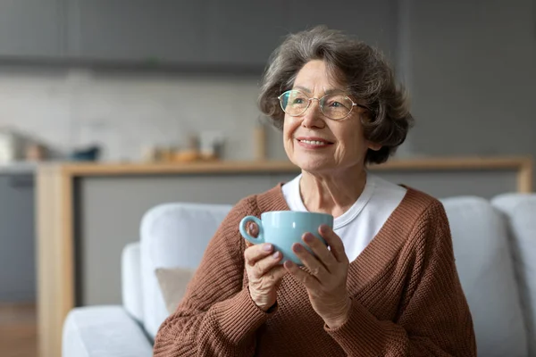 Grandmother start day with favourite beverage sitting on couch and hold in hands cup of coffee, happy retired time enjoy life concept
