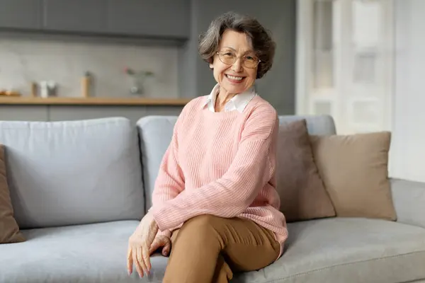 Portrait of smiling elderly woman in casual wear and glasses sitting on sofa in relaxed pose and smiling at camera, carefree senior lady in modern apartment