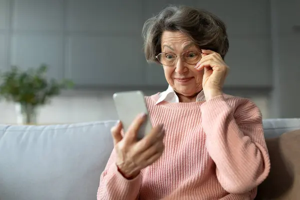 Surprised retired woman in glasses staring at smartphone, getting good news, reading text message, sitting on couch at home, free space