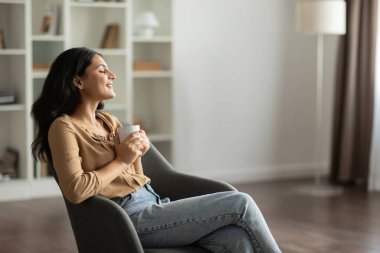 Genç Hintli bayan rahat koltuğunda oturuyor, kahve içiyor, mola veriyor ya da evde sıcak bir şeyler içiyor.