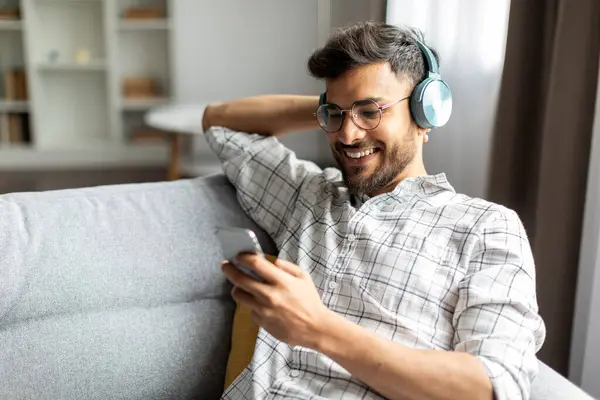 Relaxed indian guy with wireless headphones using cellphone lying on sofa, listening to music online at home, enjoying free time on weekend