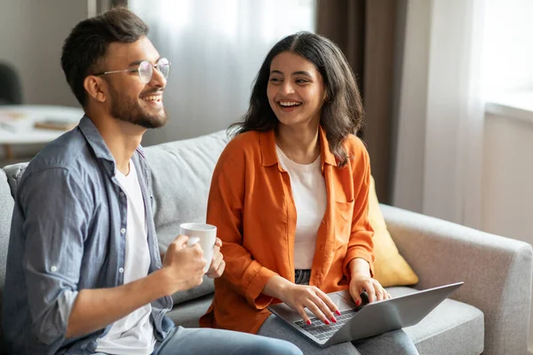 Young indian spouses using laptop and talking, choosing movie together, sitting on sofa at home, enjoying free time on weekend. Gadgets and technology