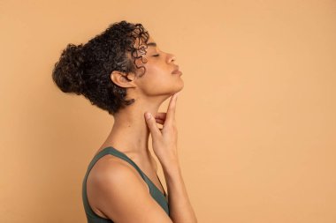 Side view of latin woman enjoying her silky skin, touching neck, empty space, beige studio background, natural beauty concept clipart