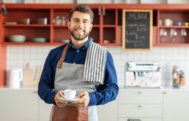 Kahve fincanı tutan ve kameraya gülümseyen bir adamın portresi. Kahve dükkanında önlüklü küçük işletme sahibi.