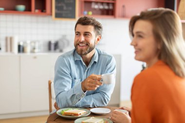 Elinde kahve fincanları olan olumlu bir adam ve kadın masanın önünde oturuyor ve ilgiyle bir tarafa bakıyor.