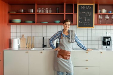 Kahve işi sahibi. Önlüklü, kahve dükkanının içindeki kameraya gülümseyen genç, güzel bayan barista.