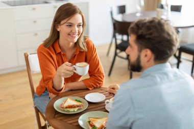 Gülümseyen kadın ve kocası kahve fincanlarıyla masanın önünde oturuyor ve içeride konuşmaktan zevk alıyorlar.