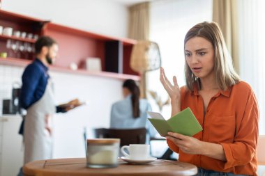 Şok olmuş yüz ifadesiyle faturasına bakan genç, şaşırmış bir kadın kahve dükkanında masada oturuyor.