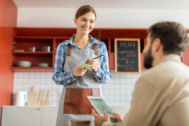 Güzel, arkadaş canlısı garson, erkek müşteriden sipariş alıyor ve not defterine yazıyor, erkeğe bakıyor ve gülümsüyor.