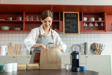 Neşeli bayan barista kahve dükkanında paket paketliyor, tezgahta duruyor ve gülümsüyor, kafede çalışmaktan keyif alıyor.