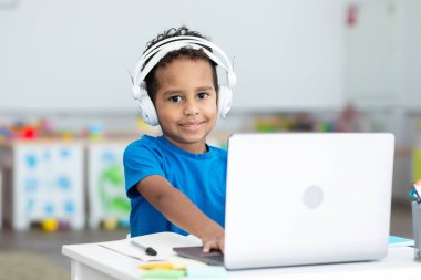 Gülümseyen siyah çocuk çevrimiçi çalışıyor, sınıfta dizüstü bilgisayar ve kulaklık kullanıyor, klavyede daktilo kullanıyor, ders alıyor.