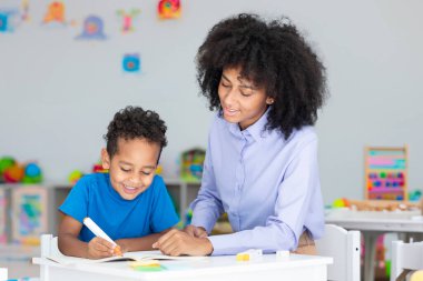 Siyahi ilkokul öğrencisiyle çalışan arkadaş canlısı bir kız öğrenci öğretmeni. Sınıfta masa başında oturmuş, kitap yazıyor.