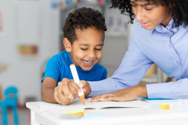 Okul öğretmeni, pozitif siyahi bir öğrenciyle çalışıyor, masada oturuyor ve sınıfın içinde yazmayı öğreniyor.