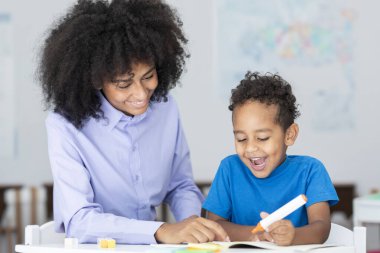 Masasında siyahi bir çocukla oturan yeni yetme öğretmen ödevlerine ve kitap yazmalarına yardım ediyor.