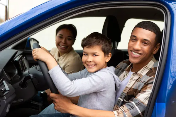 Cheerful dad teaching preteen son to drive car, boy sitting on fathers knees and smiling together to camera, family traveling by automobile