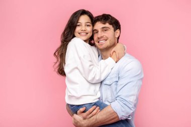 Kızına sarılıyor. Mutlu bir baba, pembe arka planda kameraya bakarken, kızına nazikçe sarılıp gülümsüyor.