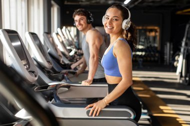 Spor giyim ve kulaklık takan neşeli bir kadın spor salonunda koşu bandı çalıştırıyor, kameraya gülümsüyor. Kaloriler kardiyo eğitimi sırasında yanar.