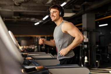 Kablosuz kulaklıklı yakışıklı adam spor salonunda koşu bandında koşuyor ve koşarken müzik dinliyor, spor yaparken eğleniyor, mekan kopyalıyor.