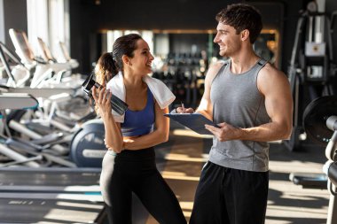Mutlu kadın elinde su şişesiyle spor salonundaki erkek antrenörüyle konuşuyor.