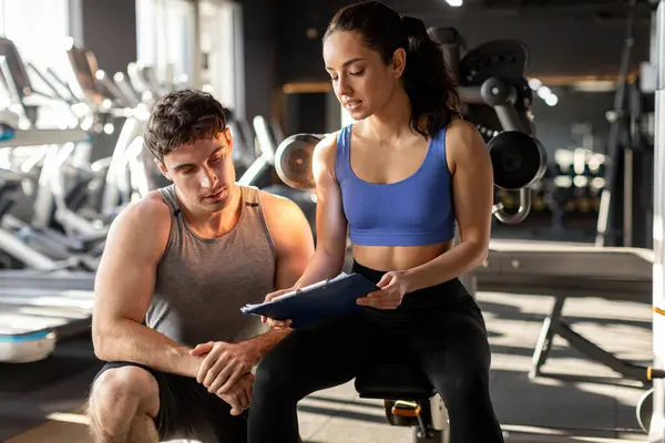 Kadın kişisel antrenör ve erkek, spor salonunda erkek öğrenciye bir şey gösteren bir panoyla fitness planını tartışıyor.