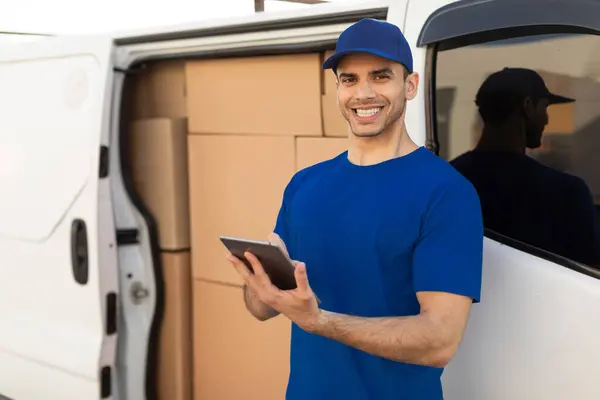 Delivery driver man smiling at camera beside his van full of parcel boxes, holding digital tablet, ready to drive to customers
