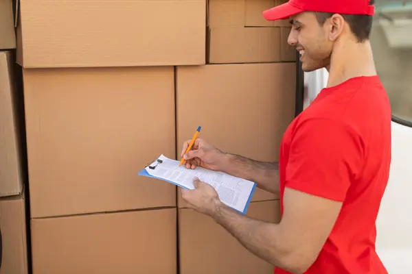 Delivery man in red t-shirt and cap checking boxes and writing in clipboard, standing next to parcels in van outdoors