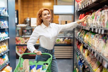 Alışveriş arabalı mutlu bayan müşteri süpermarketten gıda ürünleri alıyor, marketten yiyecek ve mısır gevreğini seçiyor, kameraya gülümsüyor.