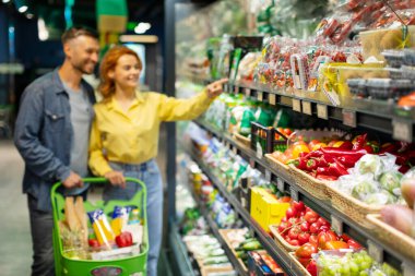 Market alışverişi. Genç çift markette sebze seçiyor, raftaki taze sebzelere odaklanıyor.