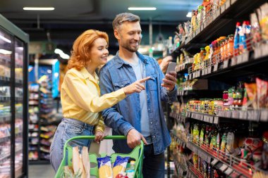 Avrupalı aile süpermarkette market alışverişi yapıyor, cep telefonu kullanıyor ve mağaza koridorunda duran ürünleri seçiyor.