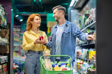 Market alışverişinde mutlu Avrupalı çift, taze sebze seçiyorlar, market arabalarıyla duruyorlar, gerekli şeyleri alıyorlar.