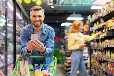 Gülümseyen adam alışveriş arabasına yaslanıyor ve akıllı telefon kullanıyor. Erkek müşteri, arka planda karısı olan marketten ürünler alıyor.