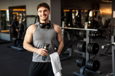 Modern spor salonunda spor sonrası idmandan sonra elinde su şişesi ve havlu tutan sporcu spor salonunda antrenman yaptıktan sonra dinlenen ve kameraya gülümseyen adam.
