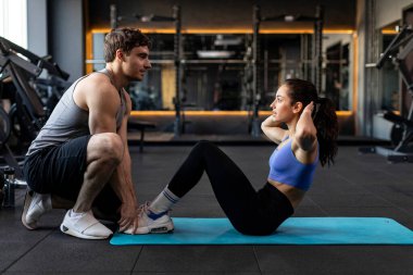 Yakışıklı erkek antrenör genç güzel bir kadına yardım ederken bir bayan spor salonunda spor minderinde, yan gözle oturuyor.