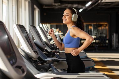 Spor salonundaki koşu bandında koşan, kulaklıkla müzik dinleyen, gülümseyen, sabah antrenmanından keyif alan genç ve güçlü bir spor kadını.