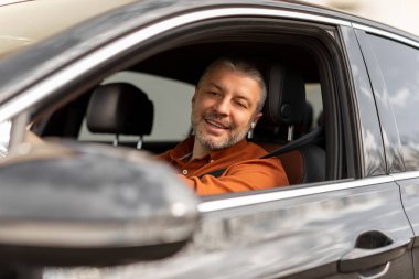 Mutlu, orta yaşlı ve araba kullanan yakışıklı adam araba yolculuğunun tadını çıkarıyor, açık pencereli aynaya bakıyor.