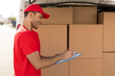 Müşterisine teslim edilmeden önce teslimat listesini kontrol eden, paketlerle dolu bir minibüsün yanında duran profesyonel bir teslimatçı.
