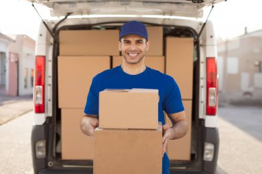 Genç Avrupalı erkek postacı elinde paket kutuları taşıyan ve gülümseyen kurye. Modern paket taşıma sistemi kavramı