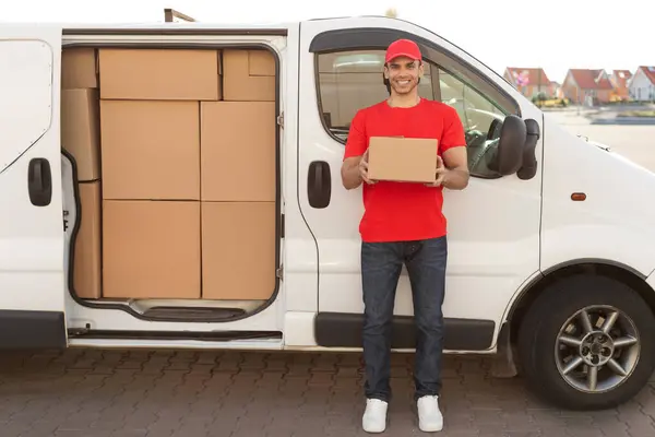 Young cheerful delivery man with parcel near car full of cardboard boxes, full length shot. Shipping service concept