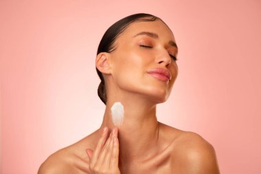 Vücuduna krem ya da losyon sürmüş, pembe stüdyo arka planında duran, doğal kozmetik ürününün reklamını yapan şehvetli genç bir kadın.