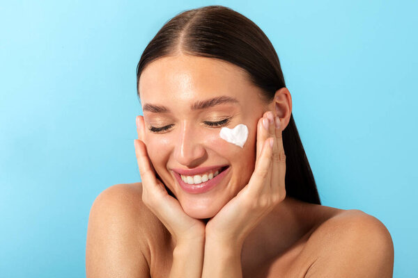 Portrait of cheerful beautiful lady with moisturizer cream in heart shape on her pretty face, recommend new beauty product, blue background