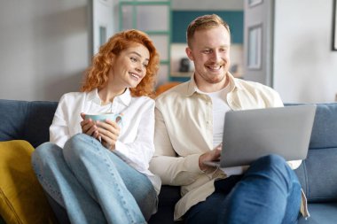 Avrupalı eşler kanepede dizüstü bilgisayarla rahatlarken ve kadın kahve içerken, çift internette sörf yaparken ve hafta sonunun keyfini çıkarırken