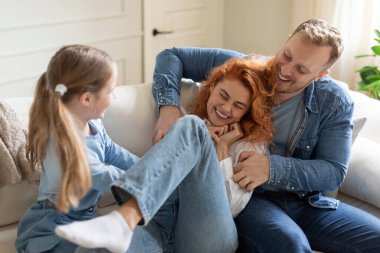 Üç kişilik neşeli bir aile evde kanepede eğleniyor, bir erkek ve bir kız annesini gıdıklıyor ve gülüyor, oturma odasında oynaşıyor.