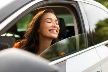 Heyecanlı genç bayan sürücü koltuğunda oturuyor ve sürüşün ve doğanın tadını çıkarırken pencereyi kapalı gözlerle dikizliyor.